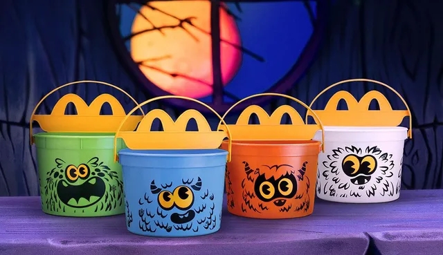 Image of McDonald's 2024 Halloween Boo Buckets showcasing four colorful designs: a classic white bucket, a vibrant orange bucket, a spooky green bucket, and the first-ever blue bucket, each adorned with playful monster faces and accompanied by stickers for decorating.